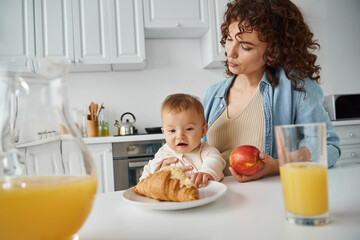 Obraz na płótnie Canvas mother and baby near croissant, ripe apple and fresh orange juice in kitchen, delicious breakfast