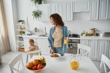 woman with orange juice and croissant near fruits and child playing with wooden tongs in kitchen