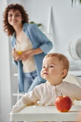 baby girl sitting near ripe apple next to mother with glass of orange juice on blurred background