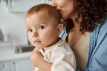 woman kissing head of cute little daughter while holding her in hands at home, blissful motherhood