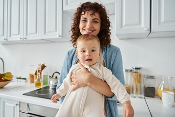 loving mother with wavy hair hugging adorable daughter in romper at home in kitchen, family bliss