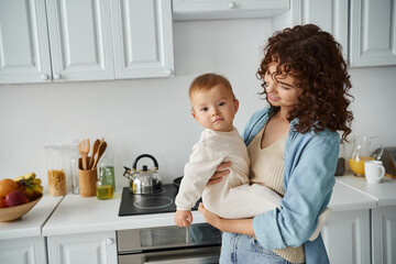 smiling curly woman holding baby girl in romper looking at camera in cozy kitchen, happy family - obrazy, fototapety, plakaty