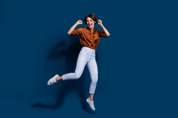 Full length photo of shiny lucky girl dressed brown shirt spectacles jumping high rising fists isolated blue color background