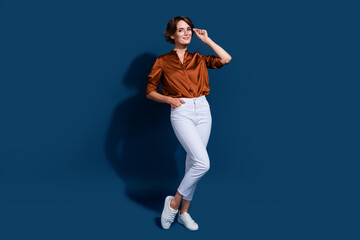 Full size photo of pretty young girl touch specs hand pocket posing wear trendy brown blouse isolated on dark blue color background