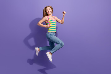Fototapeta na wymiar Full length photo of lovely teen blonde lady jump running raise fists dressed stylish striped garment isolated on violet color background