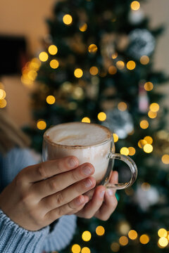 Closeup vertical cropped shot of unrecognizable little girl holding in hands mug with hot cocoa on blurred background bright bokeh lights of Christmas tree garland. Concept of home festive atmosphere.