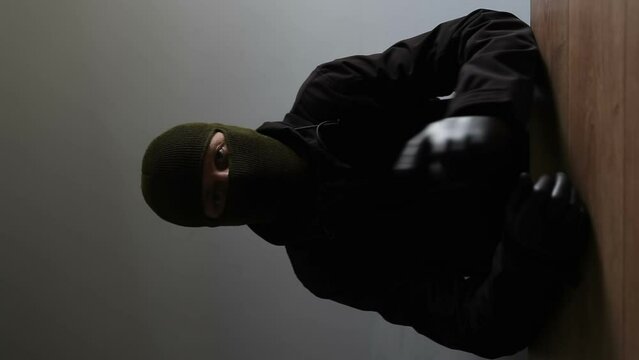 An unknown man in a black jacket and balaclava sits at a table, looks into the camera lens, speaks and sets his terms with gestures. Vertical video. Anonymous video with threats.