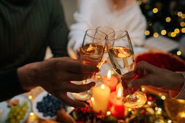 Close-up cropped shot of unrecognizable male and female friends or family clinking glasses with champagne, celebrating xmas, new year or birthday party, sitting at holiday dinner table with candles.