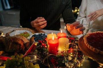 Close-up cropped shot of unrecognizable man using match lighting candles sitting at holiday table with loving family in dark living room with cozy interior, on Christmas eve, home festive atmosphere.