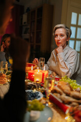 Vertica shot of unrecognizable young man holding glass with champagne, talking toast to family sitting at festive Christmas dinner table during holiday party in dark room with beautiful lighting.