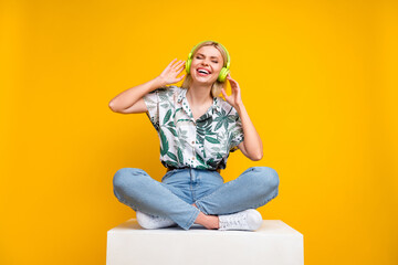 Full length photo of good mood girl dressed print blouse sit on cube listen playlist in headphones isolated on yellow color background