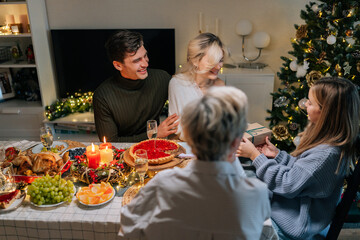Side view of happy adorable little girl receiving Christmas gift from loving family at home on xmas eve. Smiling mother giving present to daughter on holidays. Concept of festive atmosphere.