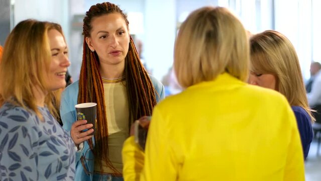 four women in the office during a break communicate drink tea talk gossip laugh yellow blue clothes celebrate a corporate party blondes. one with red dreadlocks on her head break