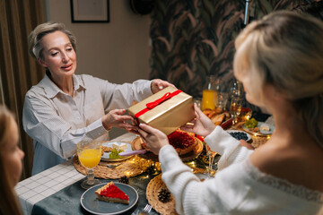 Rear view of happy young woman receiving gift from loving mother-in-law sitting with family at dinner feast table at home on Christmas Eve. Blonde female giving present to beloved daughter on holidays