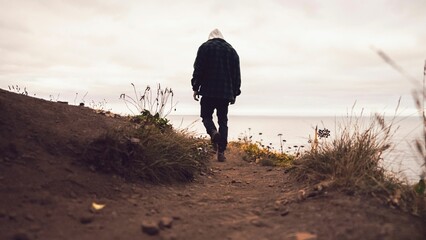 Adult male with a hoodie walking along a sandy path by the shore on a cloudy day