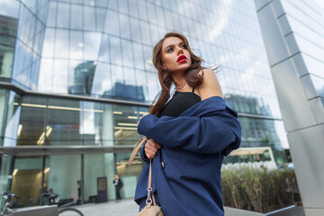 Glamorous beautiful young fashionable urban woman with hairstyle and makeup in a fashion suit with a top with a bag walking in the modern city near a glass business building