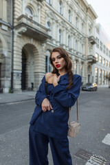 Fashion beauty elegant business girl with makeup and hairstyle in stylish blue suit with trendy bag stands and poses in the city near a vintage building. Chic pretty woman
