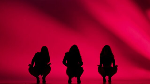 Three women silhouettes dancing heels dance in a studio. Bright red neon light, shadowed background. Black sexy costume, high heels. Modern sensual choreography. Full length.