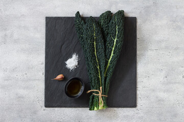 Bunch of black tuscan kale (cavolo nero or lachinato kale), olive oil, garlic and salt on a black slate board on a gray textured background, top view