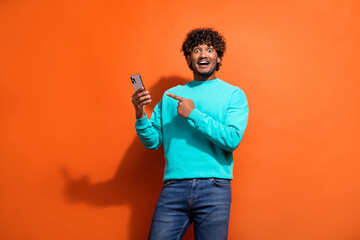 Photo of young guy with shock expression wear teal pullover pointing finger his new smartphone features isolated on orange color background