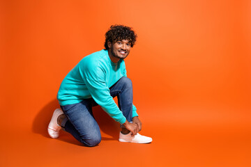 Full length body photo of young man sitting and lacing up his brand new lacoste brand sneakers isolated on orange color background
