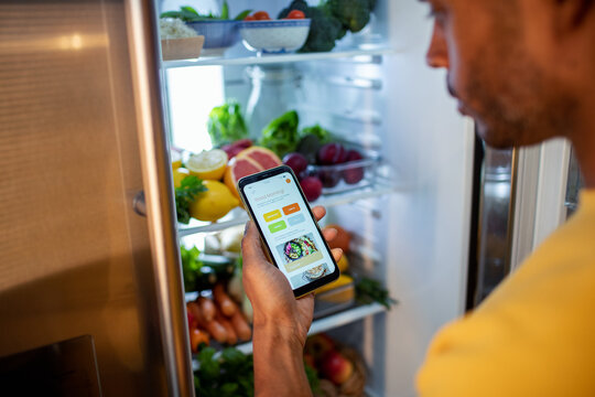 Man checking a meal planning app while looking into a refrigerator filled with fresh vegetables