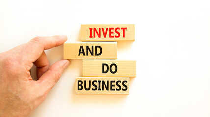 Invest and do business symbol. Concept word Invest and do business on beautiful wooden block. Beautiful white table white background. Businessman hand. Invest and do business concept. Copy space.