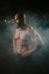 Fototapeta na wymiar A muscular bald man poses shirtless in the dark amid smoke. A bodybuilder shows off his form in the gym.