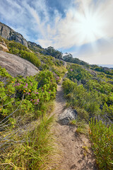 A mountain trail with cloudy blue sky and lensflare. Landscape of countryside dirt road for hiking on adventure walks along a beautiful scenic trail with lush shrubs in Cape Town, South Africa