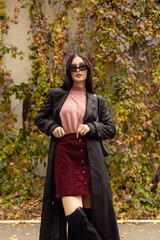 Young woman in black sunglasses, leather raincoat, pink turtleneck and red skirt. Natural. Attractive woman in park. Youth fashion