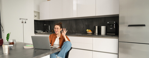 Portrait of happy friendly woman, sitting at home with laptop, waving hand at computer camera,...