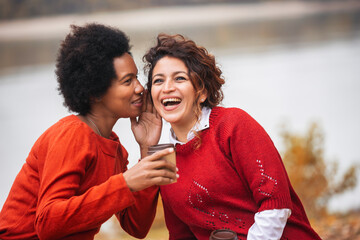 Two happy female friend talking and sharing life stories, enjoying autumn outdoors , drinks coffee. Women's friendship - obrazy, fototapety, plakaty