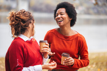 Two happy female friend talking and sharing life stories, enjoying autumn outdoors , drinks coffee. Women's friendship