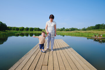Fototapeta na wymiar A child and a boy with a grandmother walk on a wooden pier by the lake. Mom and baby walk along the pier on the lake. Kid aged two years