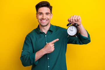 Photo of cheerful satisfied man wear green stylish clothes showing alarm clock isolated on yellow...