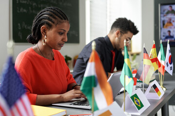 Group of international students sitting at table with flags and using gadgets at lesson in the classroom - obrazy, fototapety, plakaty