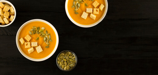 Pumpkin soup with seeds and crackers in the white bowls on the black wooden background. Top view. Copy space.
