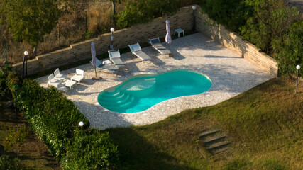 Aerial view of a swimming pool with ladder, belonging to a large villa. The pool is empty and no one is swimming. Around the water there is a stone floor.