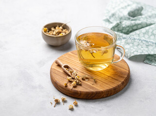 Chamomile herbal tea in a glass cup on a wooden board on a light background with dry flowers. The...