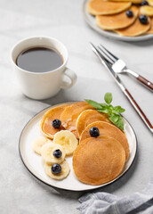 Delicious oat banana pancakes with fresh fruit slices, berries, mint and honey on a light background with a cup of black coffee.
