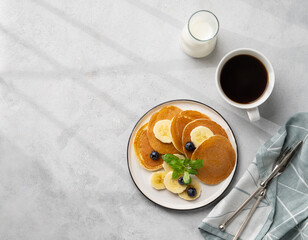 Delicious banana pancakes with fresh fruit slices, berries, mint and honey on a light background with a cup of black coffee, soy milk and shadow. Healthy vegetarian diet breakfast.