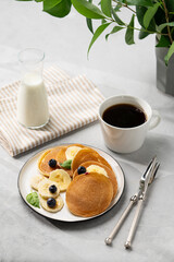 Delicious banana pancakes with fresh fruit slices, berries, mint and honey on a light background with a cup of black coffee, soy milk and green branch.
