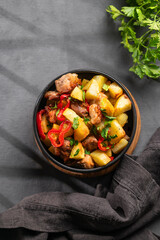 Fried pork meat with potatoes, onions and peppers. National homemade Georgian dish ojakhuri in a black frying pan on a dark background with fresh herb and shadows.