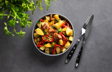 Fried pork meat with potatoes, onions and peppers. National homemade Georgian dish ojakhuri in a bowl on a dark background with fresh herb, fork and knife.