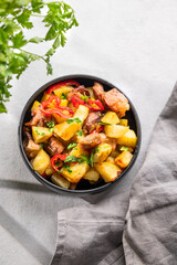 Fried pork meat with potatoes, onions and peppers. National homemade Georgian dish ojakhuri in a black bowl on a light background with fresh herb, napkin and shadows.
