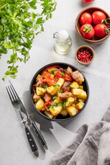 Fried pork meat with potatoes, onions and peppers. National homemade Georgian dish ojakhuri in a black bowl on a light background with fresh herb, tomato and spices.