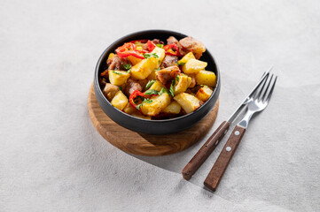 Fried pork meat with potatoes, onions and peppers. National homemade Georgian dish ojakhuri in a black bowl on a light background with shadows fork and knife.