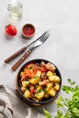 Fried pork meat with potatoes, onions and peppers. National homemade Georgian dish ojakhuri in a black bowl on a light background with fresh herb, spices and tomato.