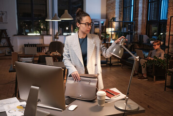Serious young mixed race businesswoman in glasses standing with bag at table and turning off light...