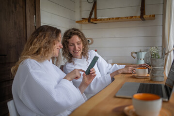 Obraz premium A woman shows her friend a funny news or photo on her smartphone while sitting at her laptop desk. Women relax in the hotel in white coats.